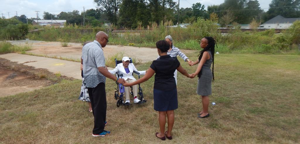 Mamie Kirkland, in wheelchair, holds hands in prayer with family and EJI staff at site of lynching