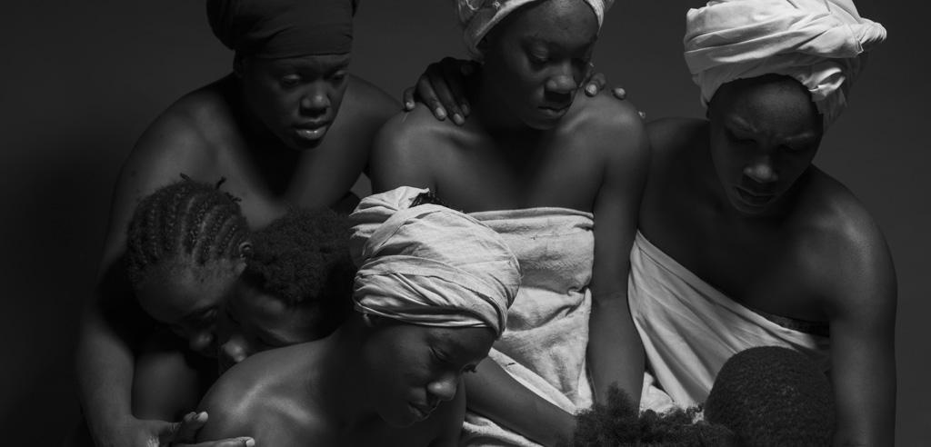 Black-and-white image of black women dressed as enslaved women huddle together with children.